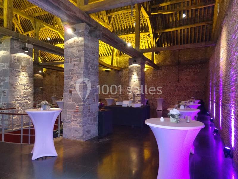Location salle Anhée (Namur) - Ferme de l'Abbaye de Moulins #34 Salle en pierre avec poutres apparentes, tables hautes blanches et éclairage violet créant une ambiance chaleureuse.