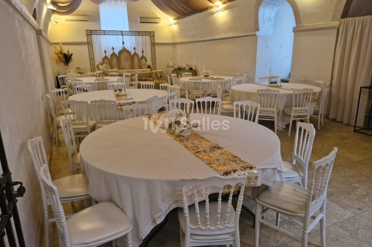 Salle de réception avec tables rondes dressées de nappes blanches et décorations dorées, éclairage tamisé.