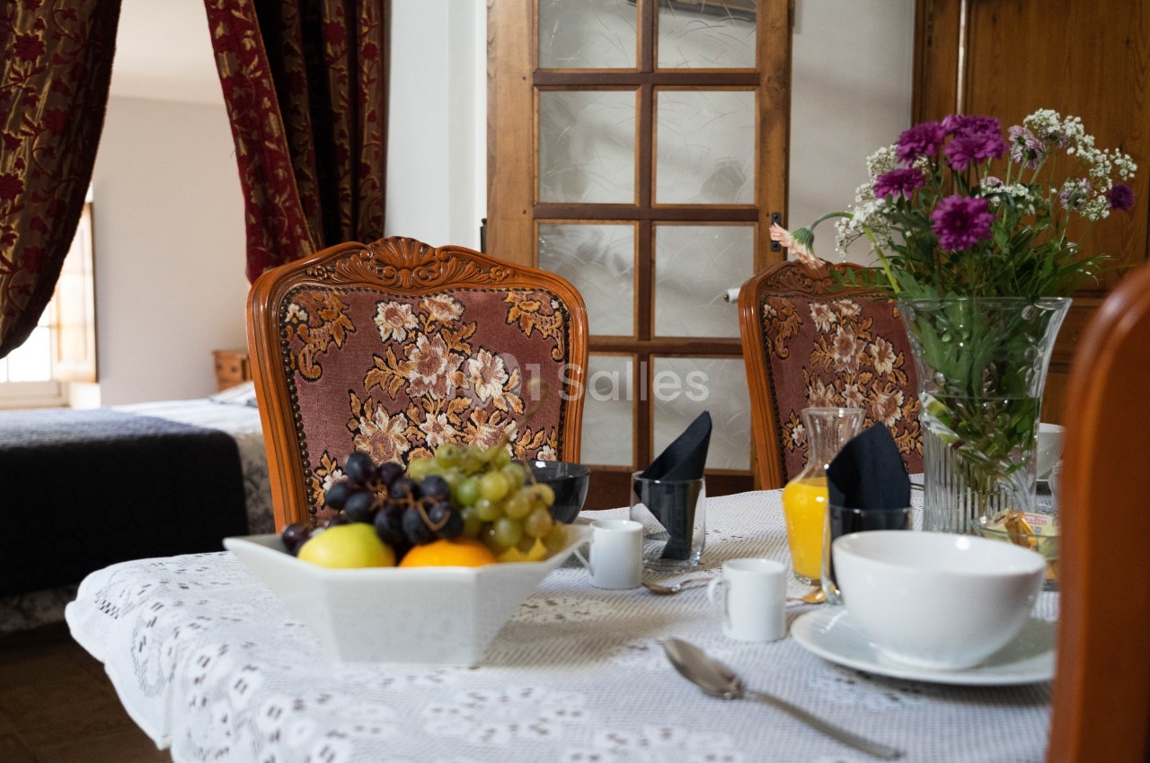 Table dressée avec une nappe blanche, contenant un bol de fruits, des fleurs, des tasses et un verre de jus d'orange.