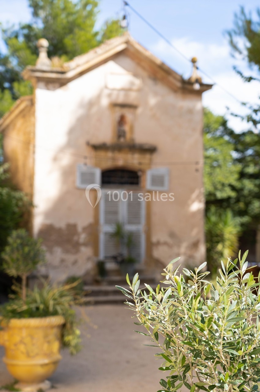 Façade d'une petite chapelle ancienne avec des volets blancs, entourée de végétation sous un ciel dégagé.