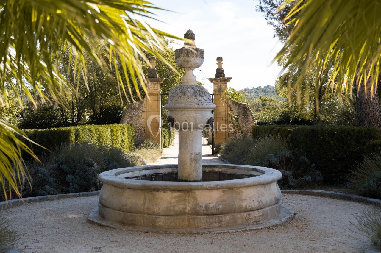 Fontaine en pierre entourée de végétation, avec une allée menant à un portail en arrière-plan.