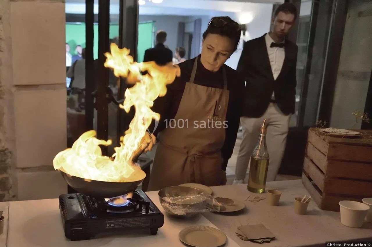 Une femme cuisine en faisant flamber un plat dans une poêle sur un réchaud lors d'un événement en intérieur.