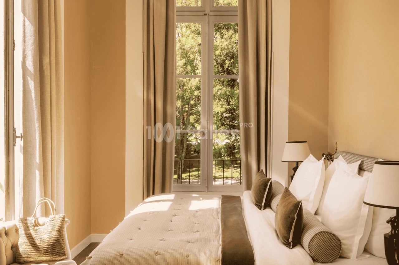 Chambre lumineuse avec un grand lit, des coussins, des rideaux beiges et une fenêtre donnant sur un jardin verdoyant.