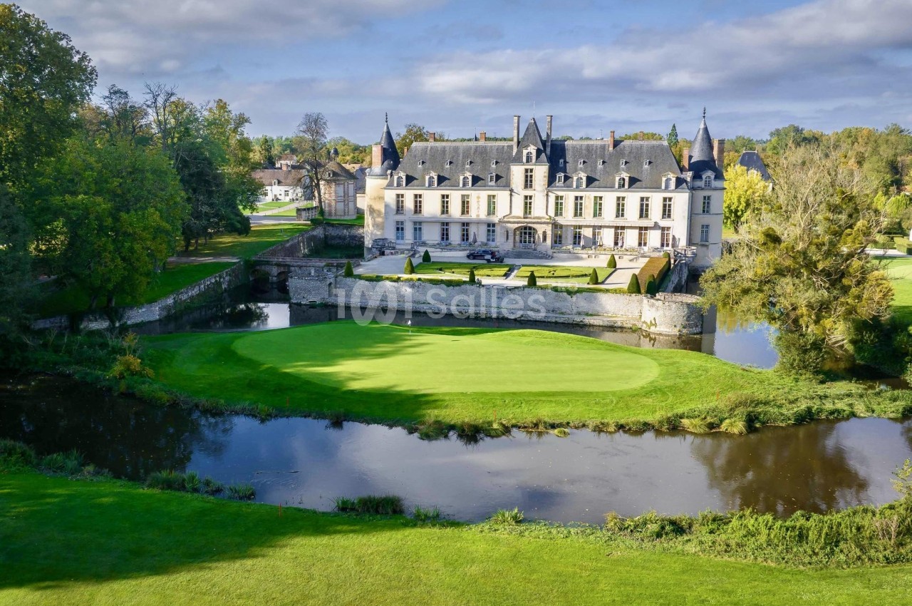 Château entouré de jardins verdoyants, bordé par une rivière sous un ciel partiellement nuageux.