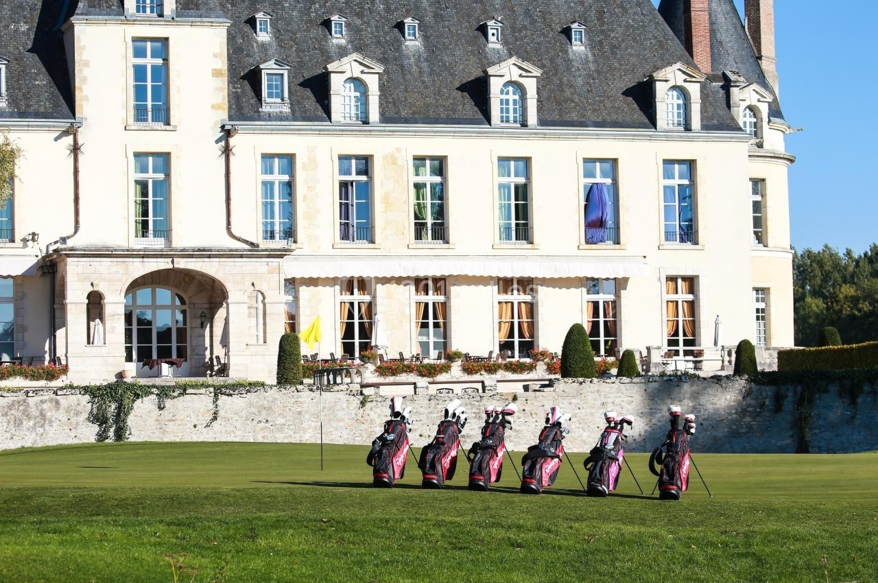 Alignement de sacs de golf sur une pelouse devant un château en pierre avec des fenêtres et des toits en ardoise.