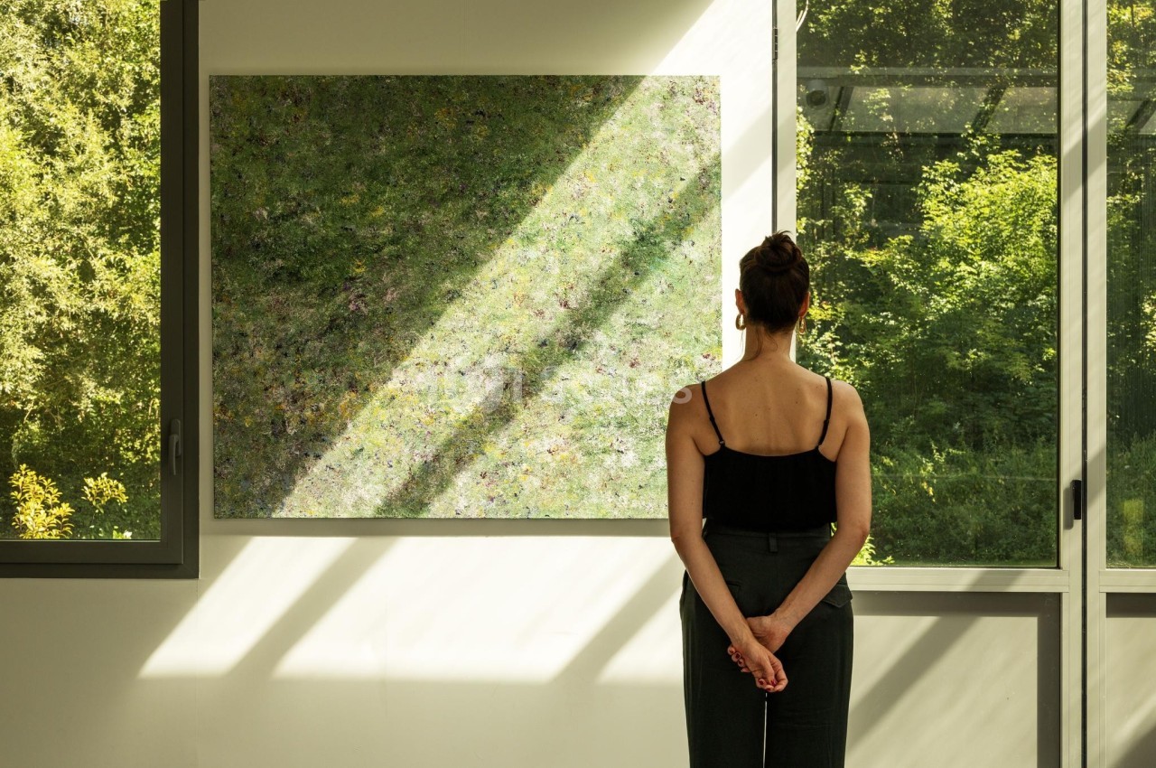 Une femme observe un tableau abstrait dans une pièce lumineuse avec de grandes fenêtres donnant sur un jardin verdoyant.