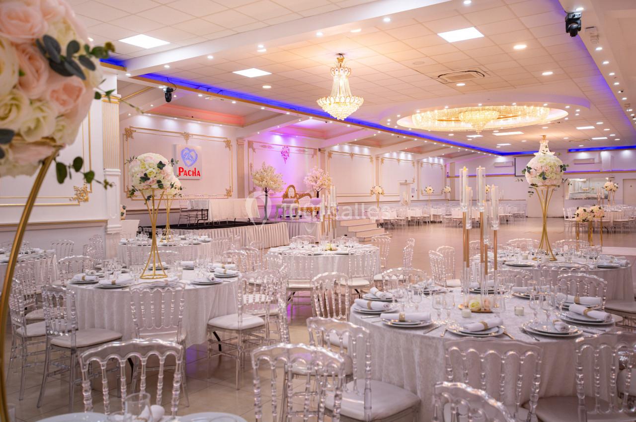Salle de réception élégante avec tables rondes dressées, chaises transparentes et décorations florales raffinées.