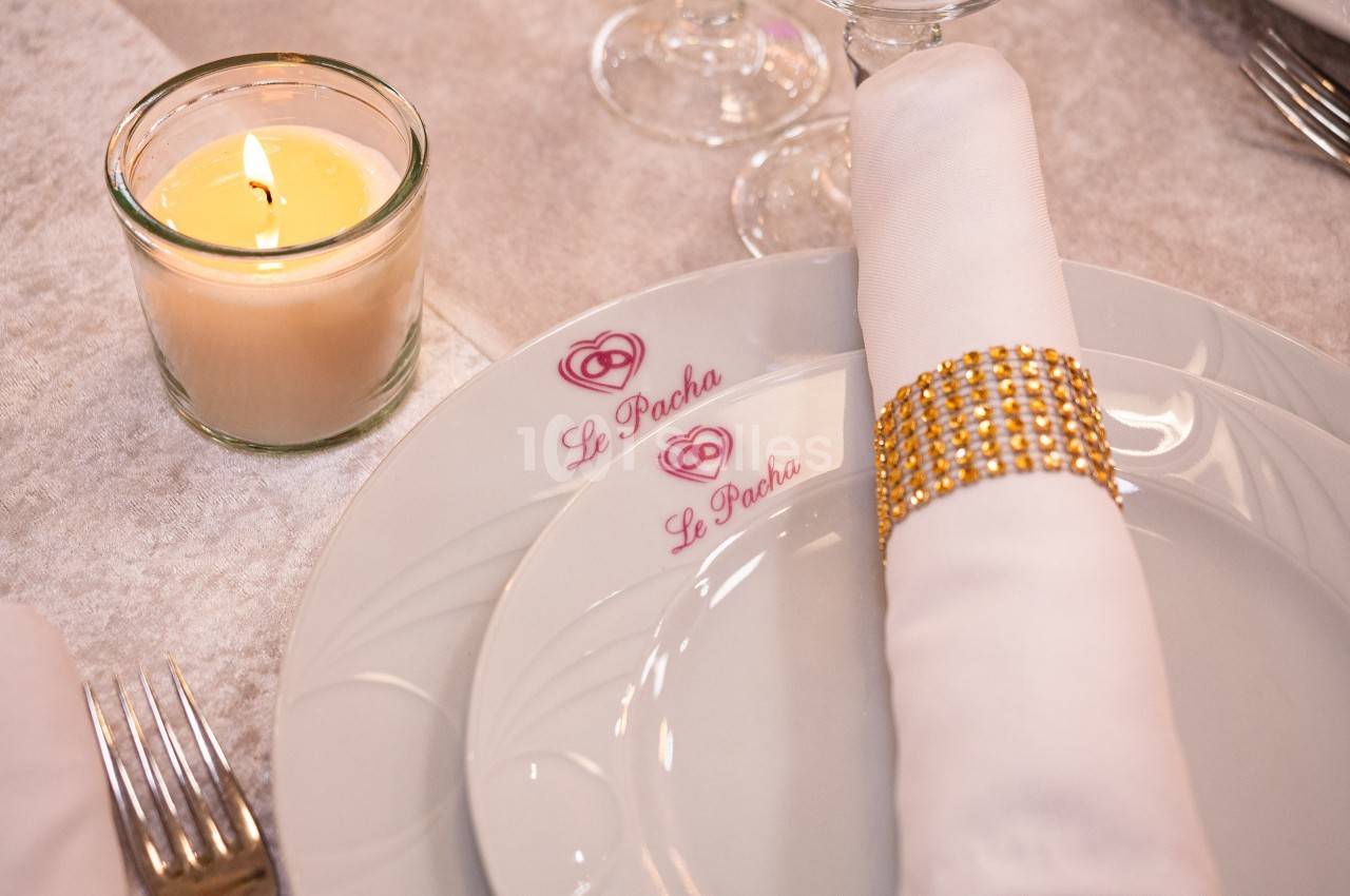 Serviette blanche ornée d'un anneau doré posée sur une assiette marquée ’Le Pacha’, avec une bougie allumée à côté.