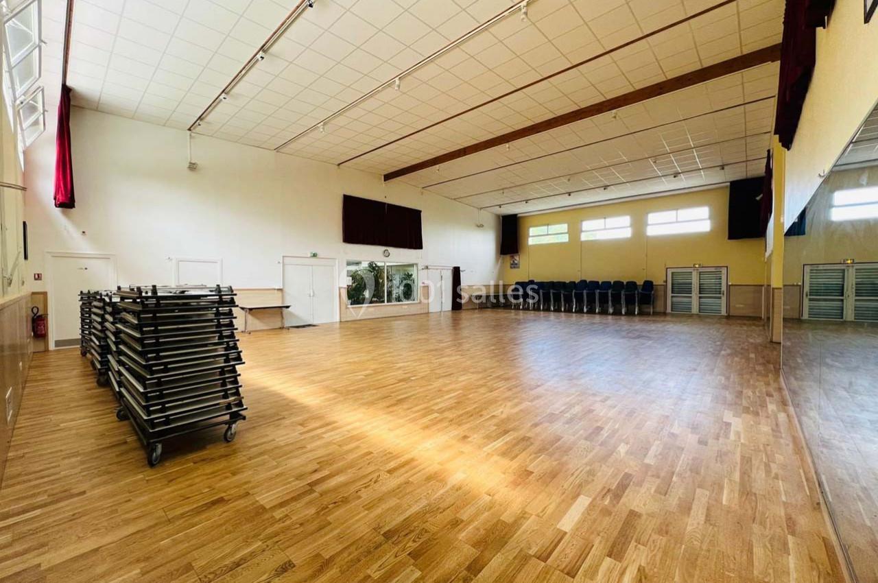 Location salle Nanteuil-lès-Meaux (Seine-et-Marne) - Centre D #18 Salle spacieuse avec parquet, chaises empilées sur le côté et tables rangées sur des chariots, éclairée par des fenêtres.