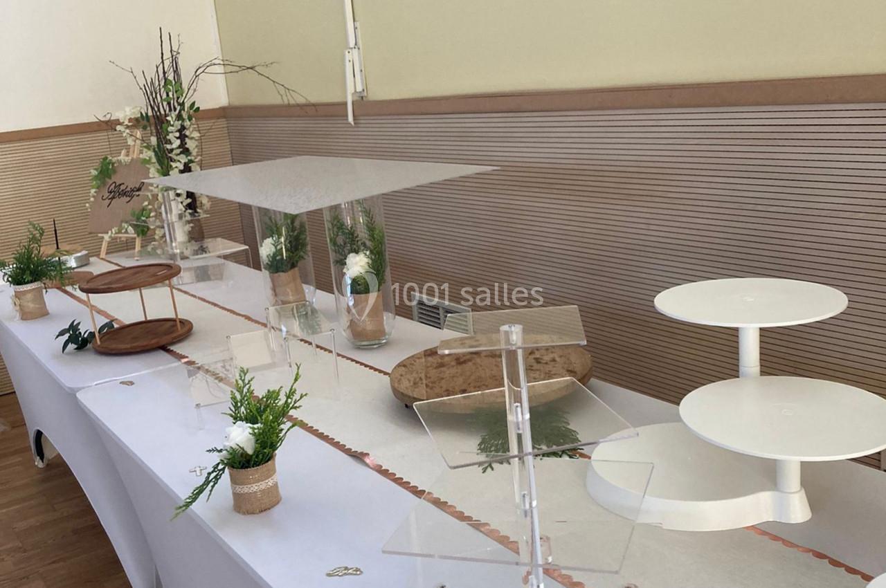Location salle Nanteuil-lès-Meaux (Seine-et-Marne) - Centre D #16 Table décorée pour un buffet, avec supports vides, plantes en pot, branches et nappes blanches dans une salle lumineuse.