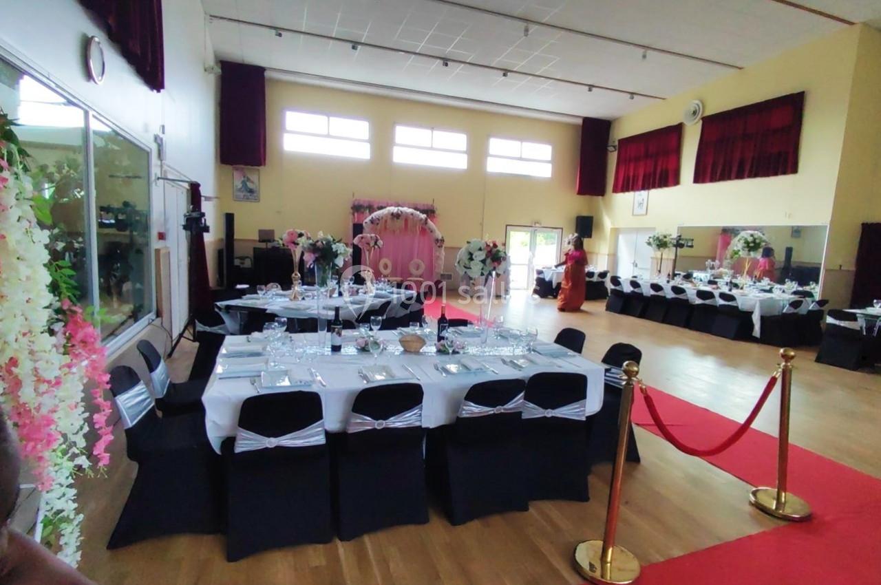 Location salle Nanteuil-lès-Meaux (Seine-et-Marne) - Centre D #1 Salle décorée pour un événement avec tables dressées, chaises noires, fleurs et tapis rouge menant à une arche florale.