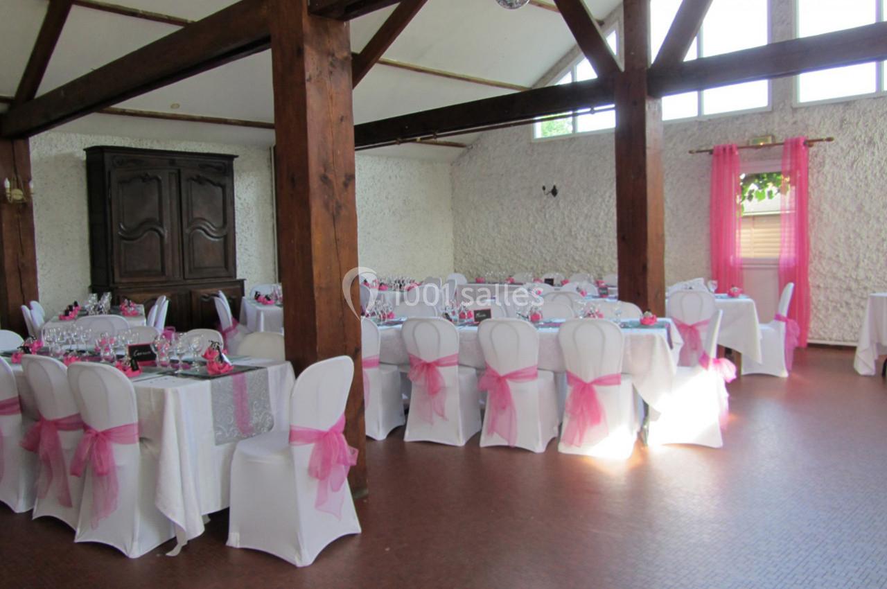 Salle de réception décorée avec des nappes blanches et des chaises ornées de nœuds roses, éclairée par la lumière naturelle.