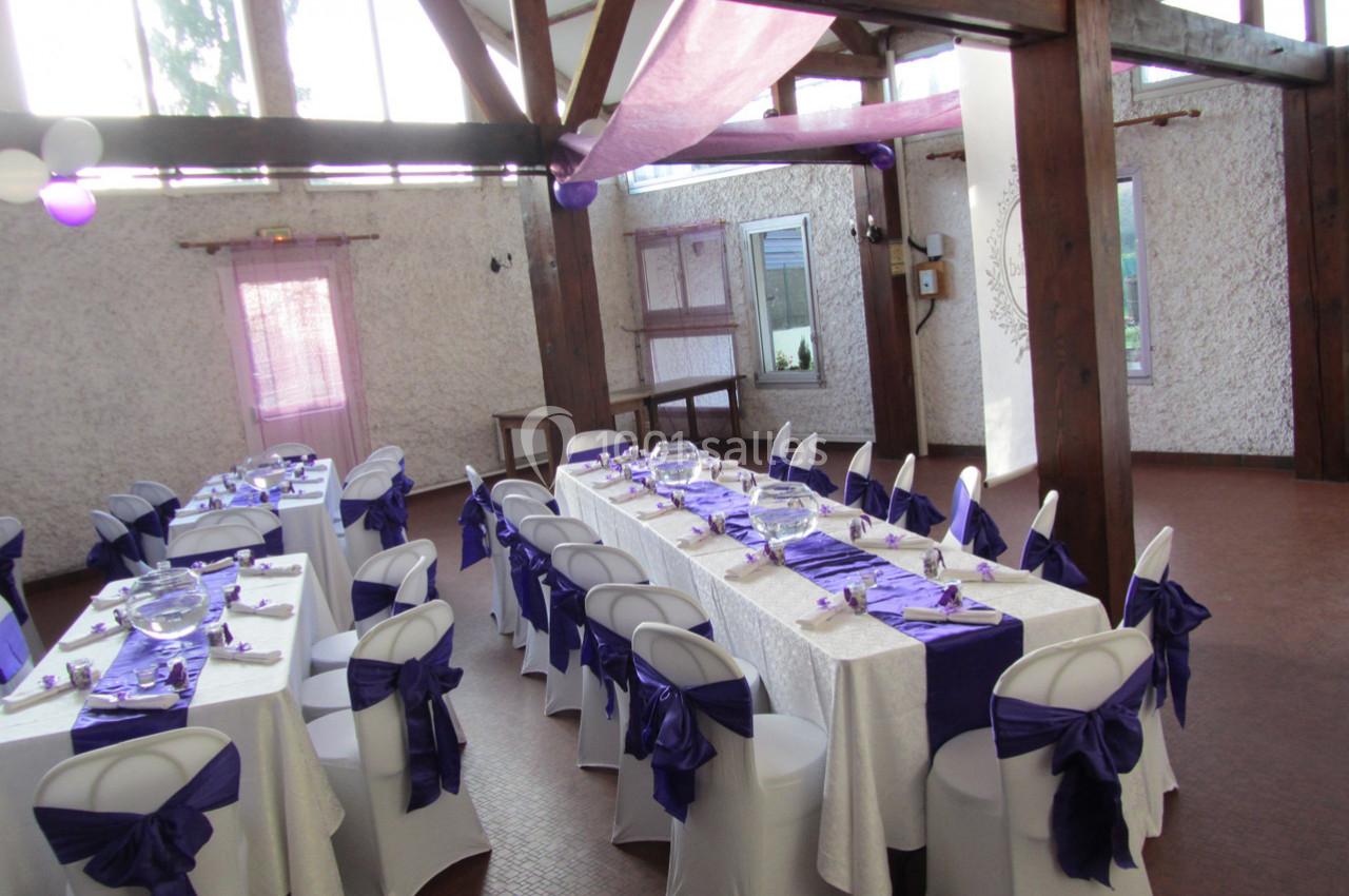 Salle décorée pour un événement, avec des tables dressées et des chaises ornées de nœuds violets.