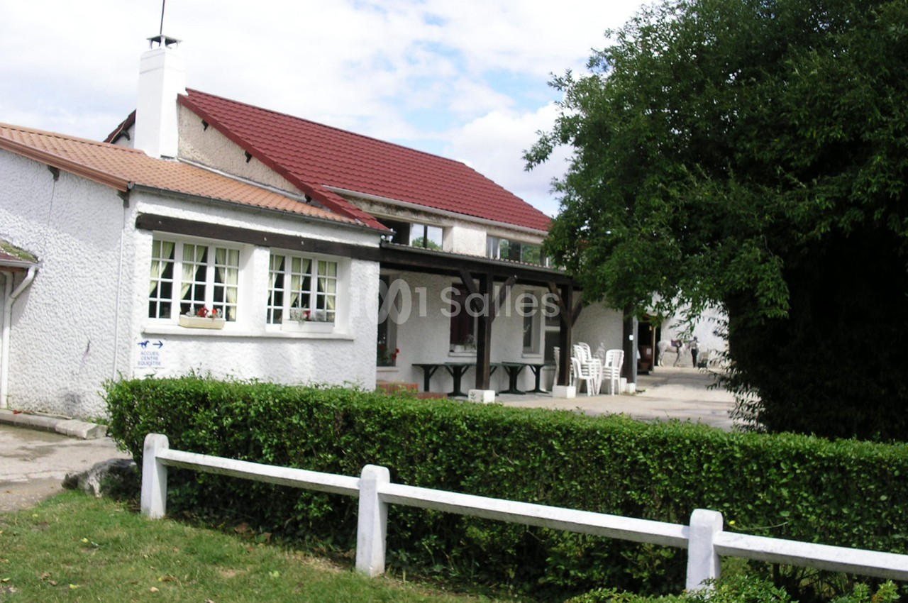 Maison avec façade blanche, toit rouge et terrasse équipée de tables et chaises, entourée de verdure et d'une haie.