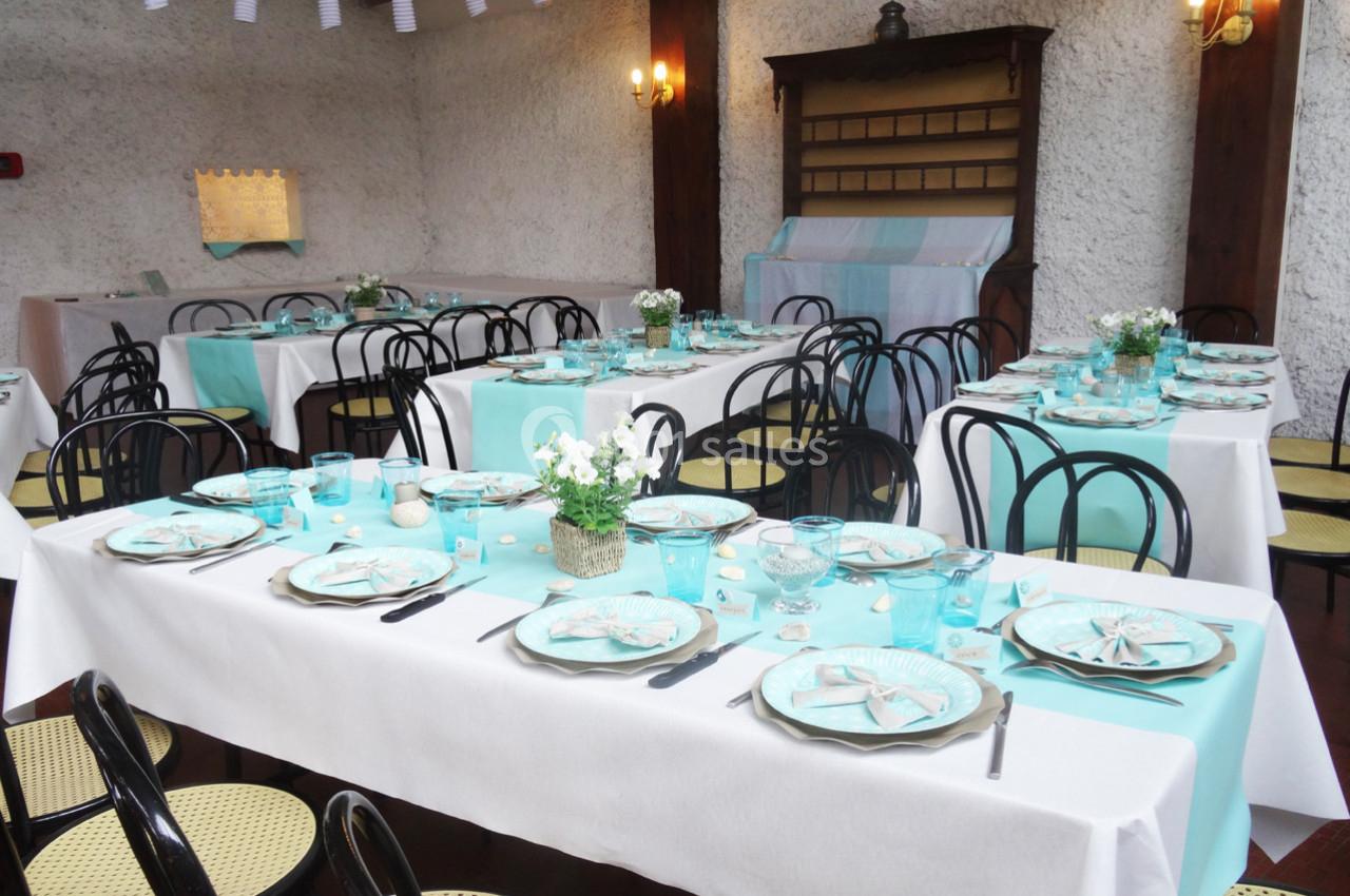 Salle décorée pour un repas, avec des tables dressées en nappes blanches et accents turquoise, chaises noires et fleurs en…