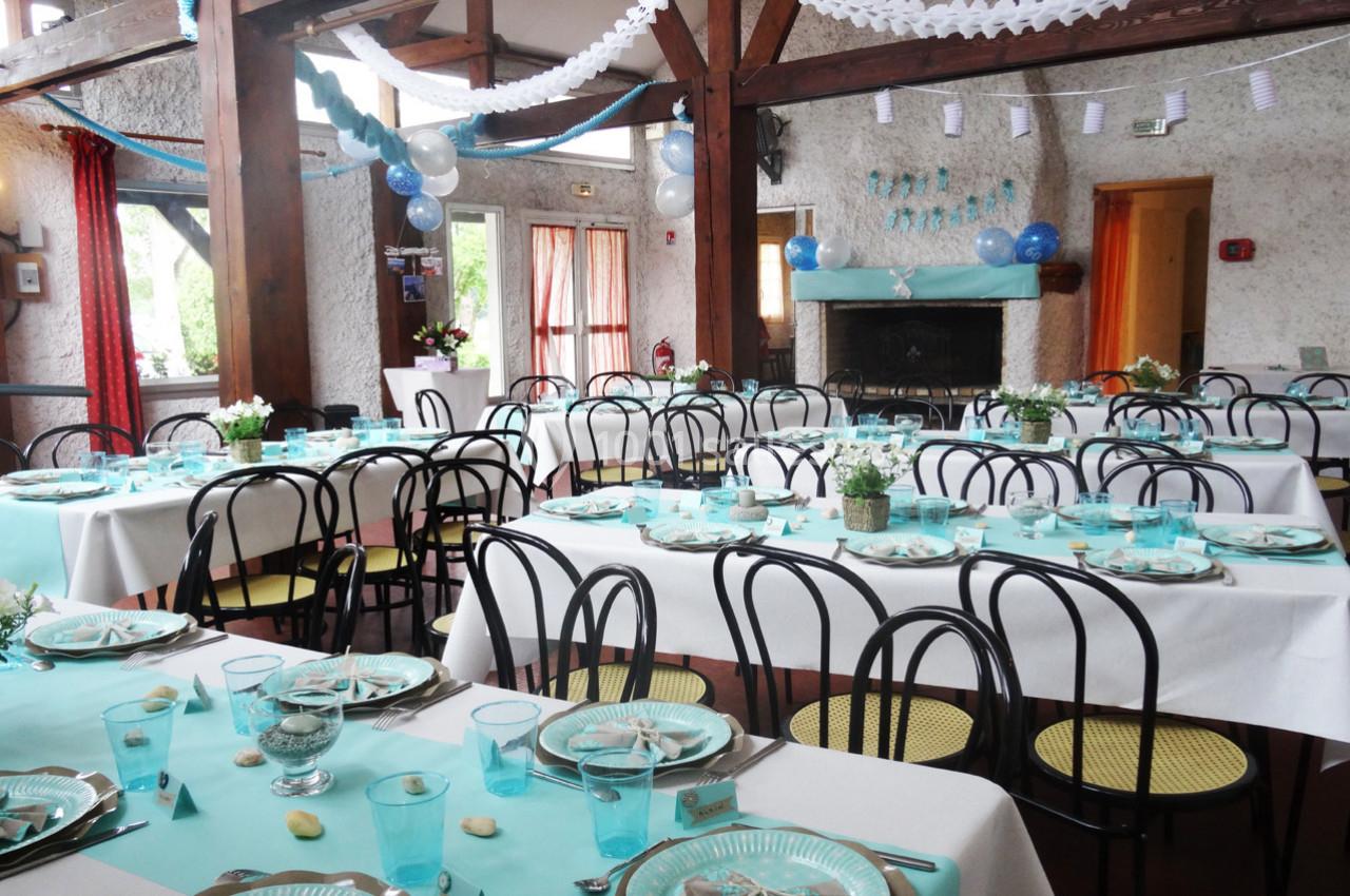 Salle décorée pour une réception avec tables dressées, nappes blanches et bleues, ballons et guirlandes suspendues.