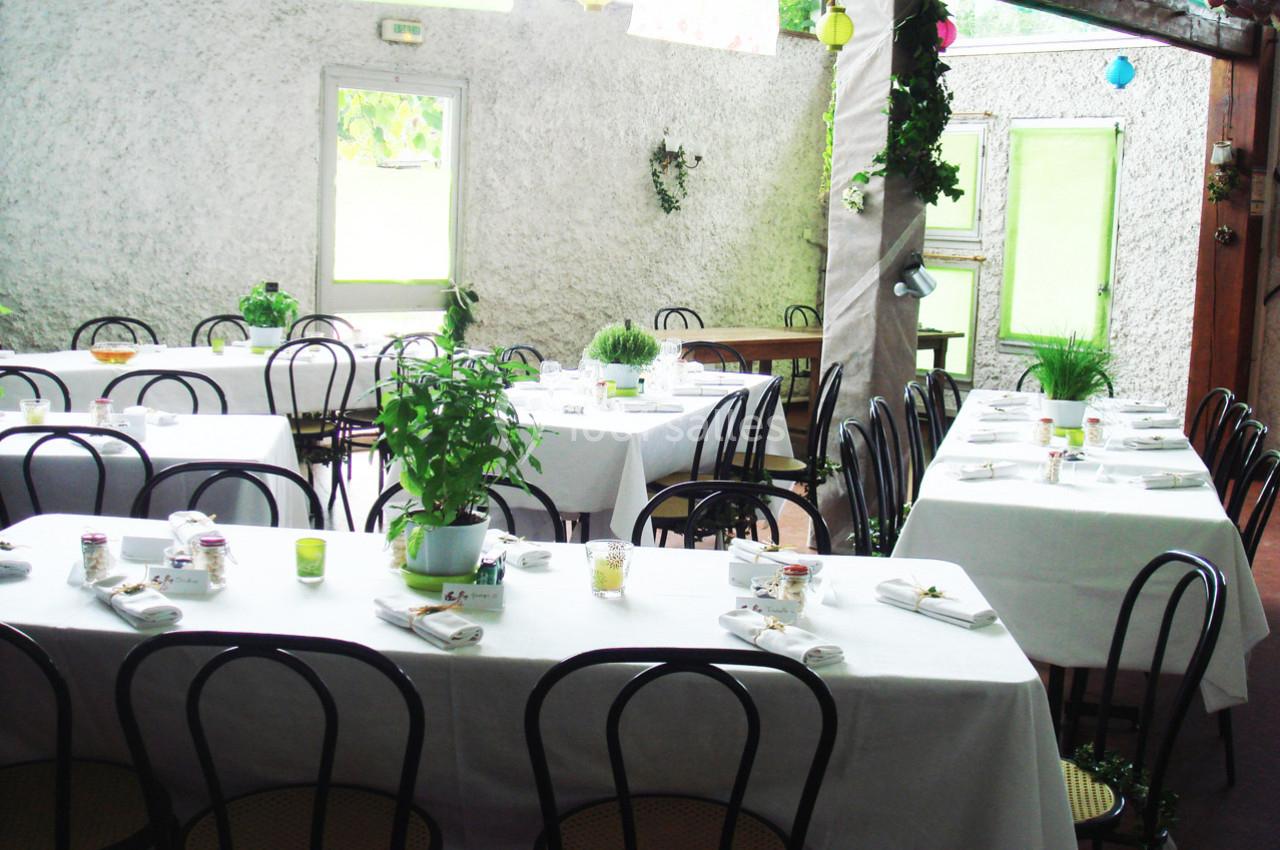 Salle lumineuse avec des tables dressées, nappes blanches, chaises noires et plantes décoratives.