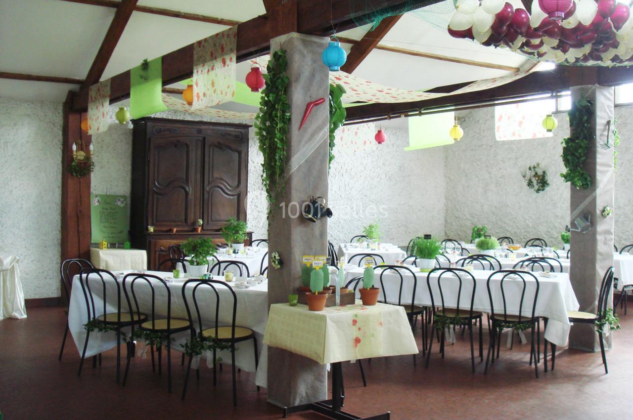 Salle décorée pour un événement, avec des tables dressées, des plantes en pot et des lampions suspendus.
