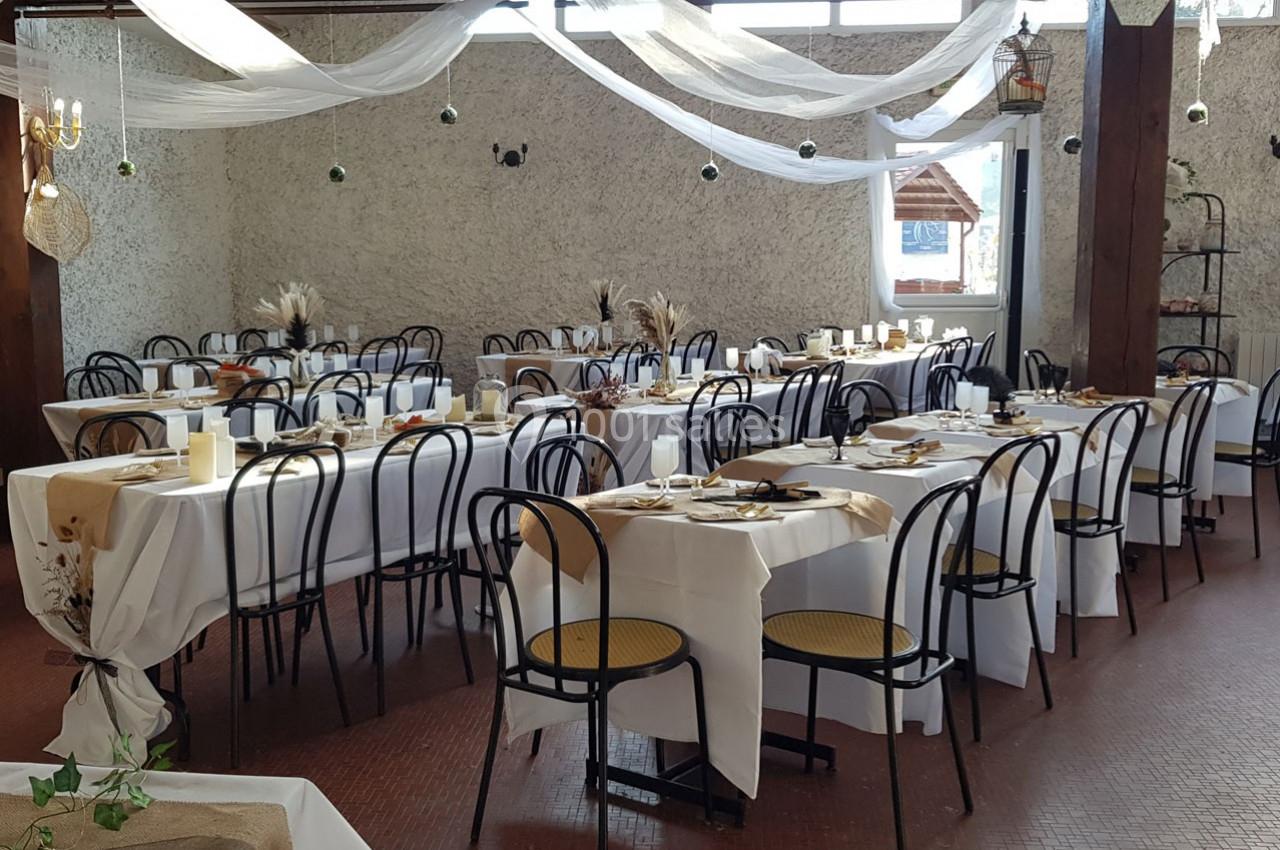Salle de réception décorée avec des tables dressées, nappes blanches, chaises noires et éléments de décoration naturels.