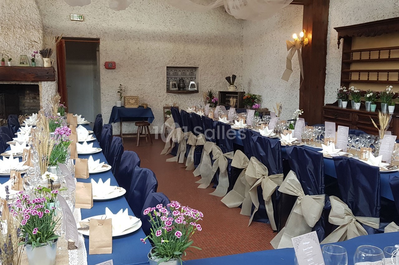 Salle décorée pour un repas festif avec tables alignées, nappes bleues, chaises ornées de nœuds beiges et fleurs en centre…