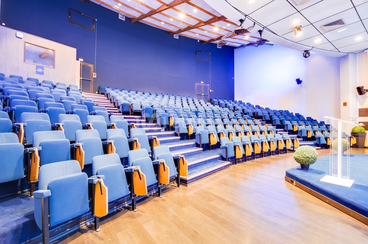 Salle de conférence moderne avec des rangées de sièges bleus vides et un podium sur une scène en bois.