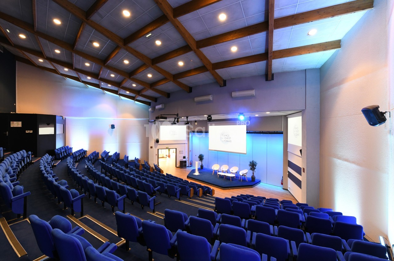 Salle de conférence moderne avec sièges bleus, scène équipée de chaises blanches et éclairage lumineux.