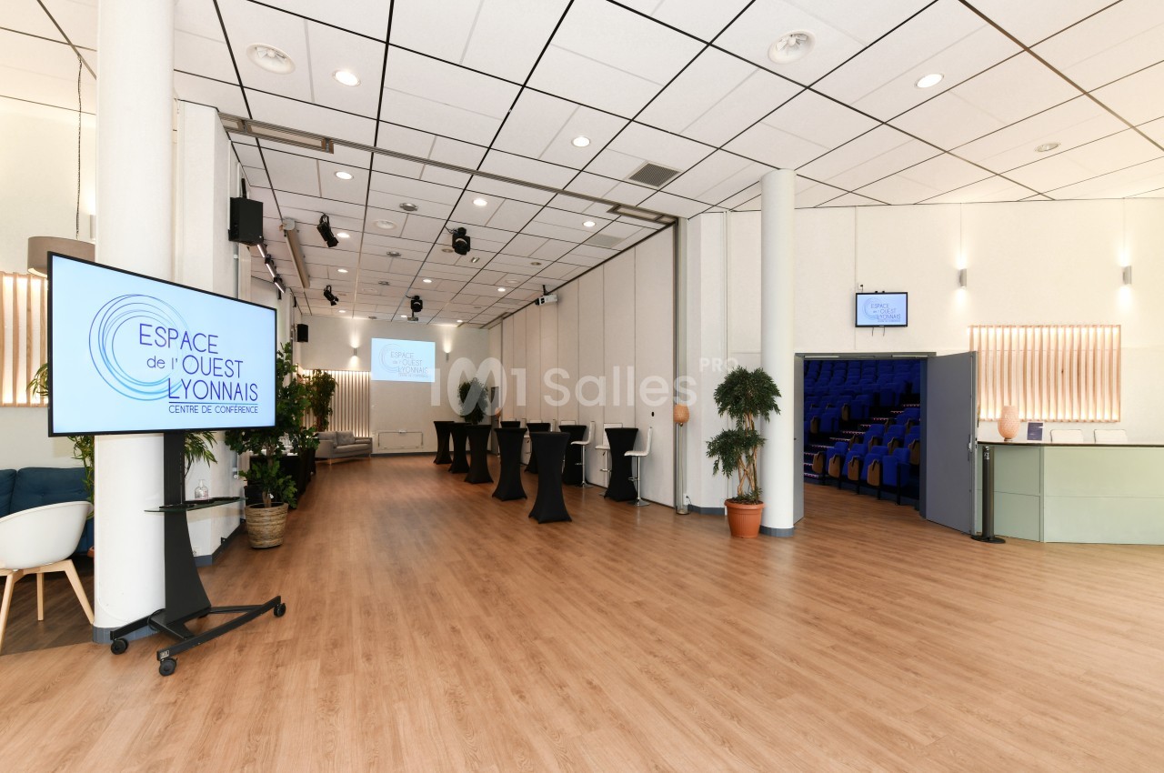 Salle de conférence lumineuse avec parquet, écrans, plantes, tables hautes et rangées de sièges visibles au fond.