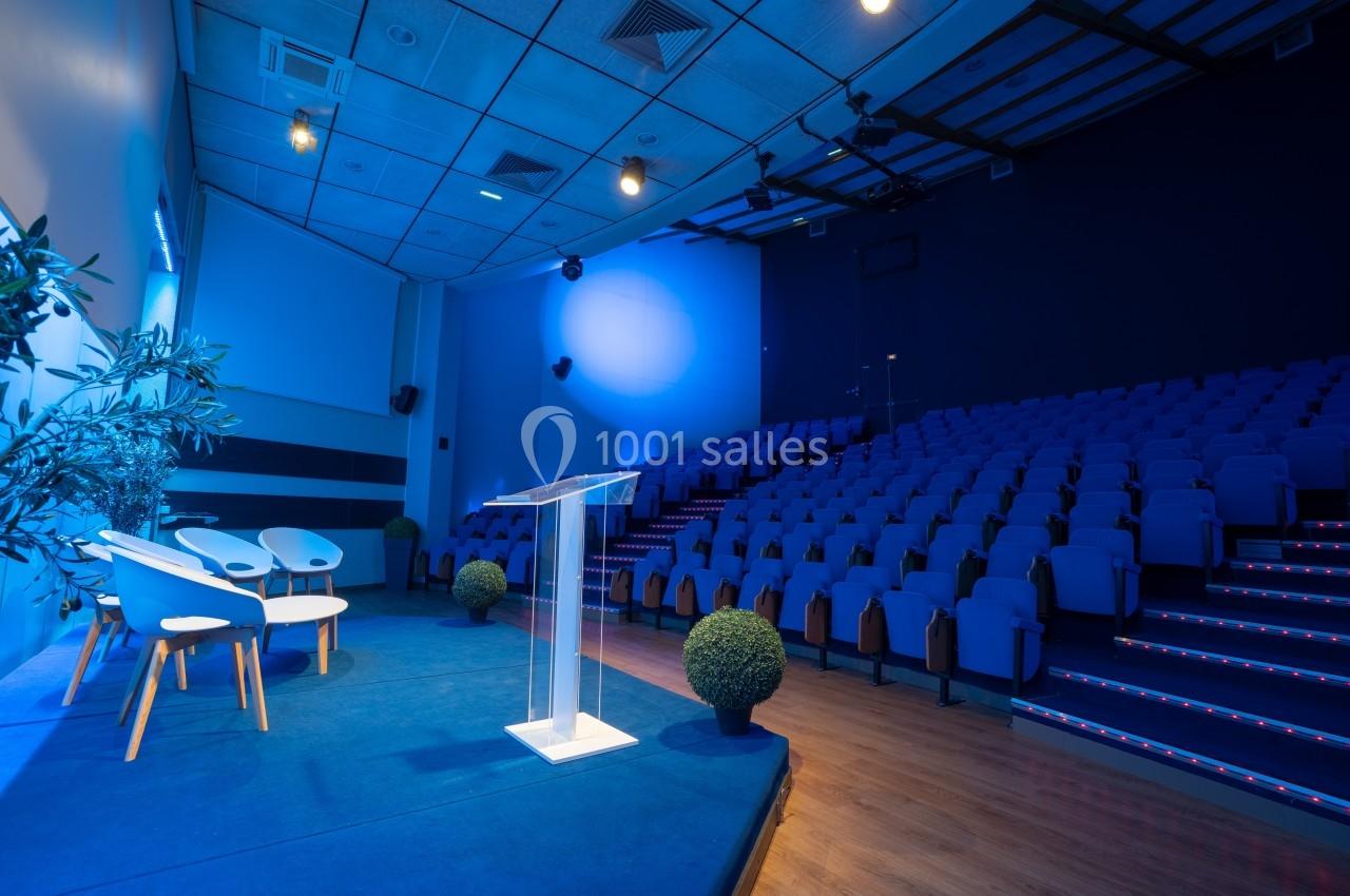 Salle de conférence vide avec un pupitre transparent, des sièges bleus en gradins et un éclairage tamisé.