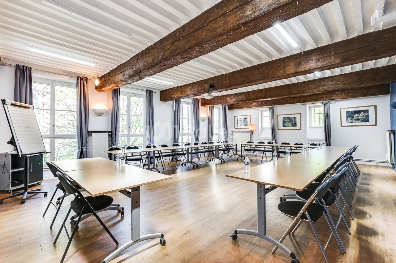 Salle de réunion lumineuse avec poutres apparentes, tables en U, chaises pliantes et grandes fenêtres donnant sur l…
