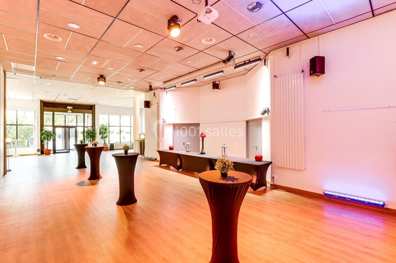 Salle lumineuse avec parquet, tables hautes drapées et décorations florales, vue sur des baies vitrées.