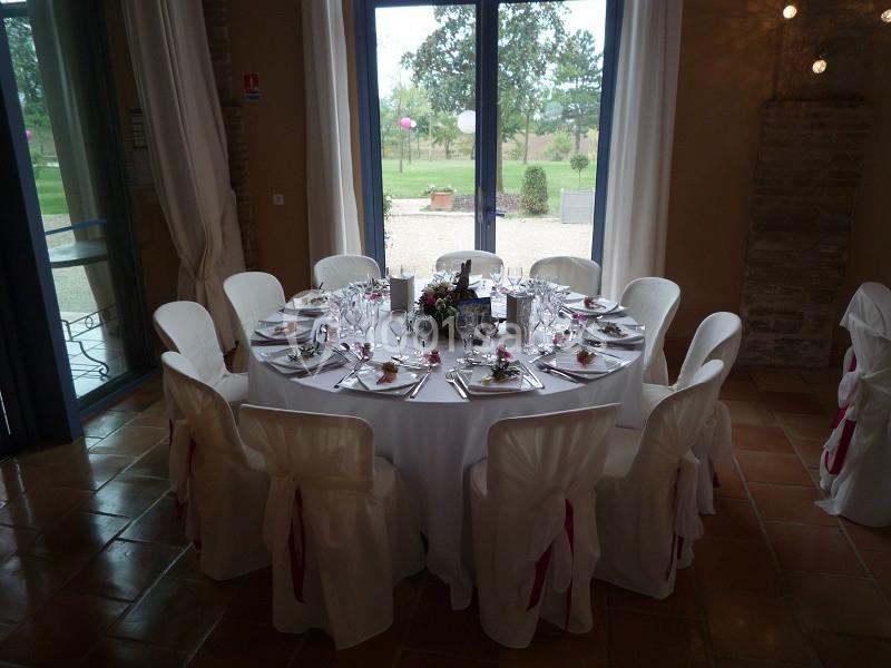 Table ronde dressée pour un événement, avec nappes blanches, chaises décorées et vue sur un jardin à travers des baies…