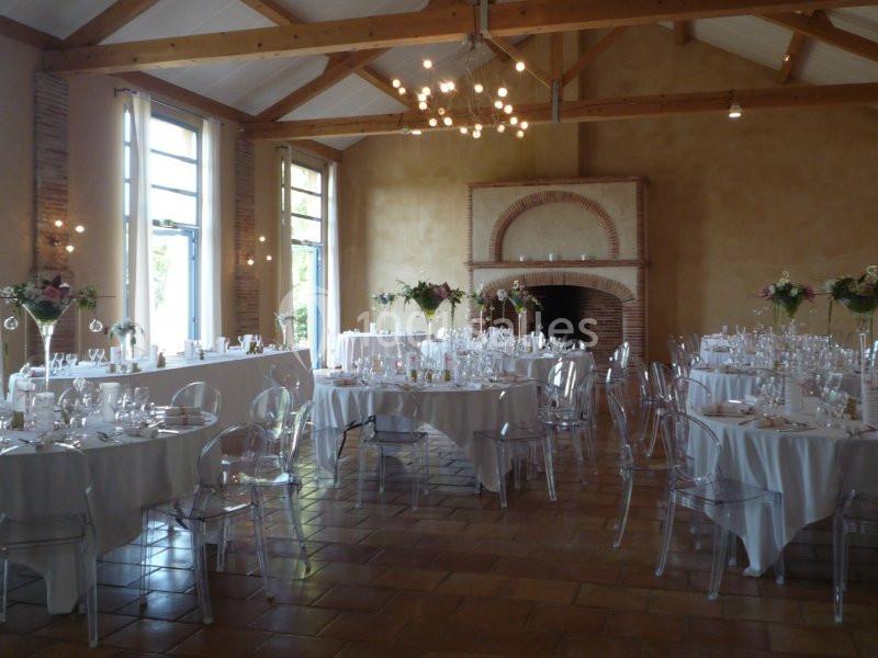 Salle de réception lumineuse avec tables rondes dressées, chaises transparentes et cheminée en briques.