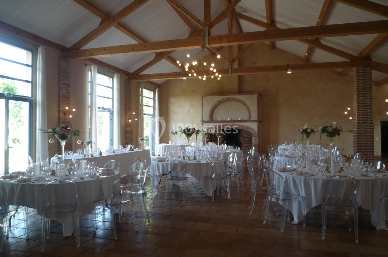 Salle de réception lumineuse avec tables rondes dressées, chaises transparentes et poutres apparentes au plafond.