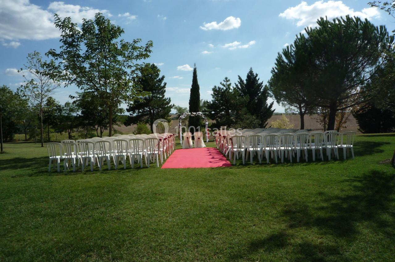 Allée centrale avec tapis rouge entourée de chaises blanches, menant à une arche décorée dans un jardin ensoleillé.