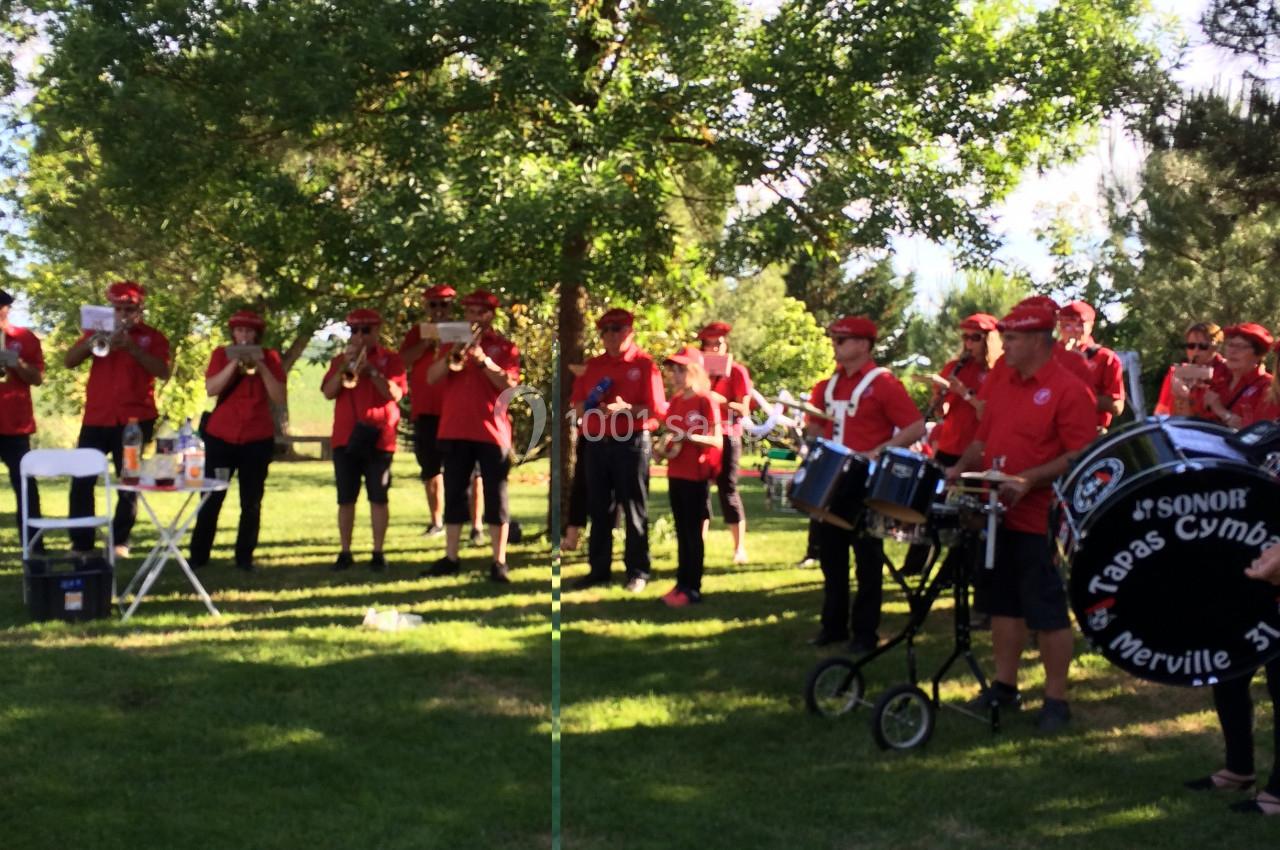 Un groupe de musiciens en tenue rouge joue dans un parc verdoyant sous un ciel ensoleillé.