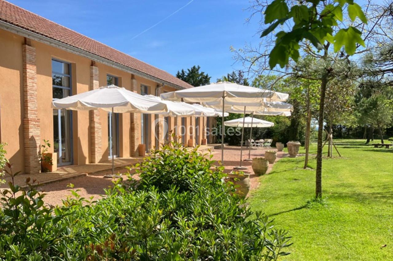 Terrasse ensoleillée avec parasols blancs devant un bâtiment ocre, entourée de verdure et d'arbres.