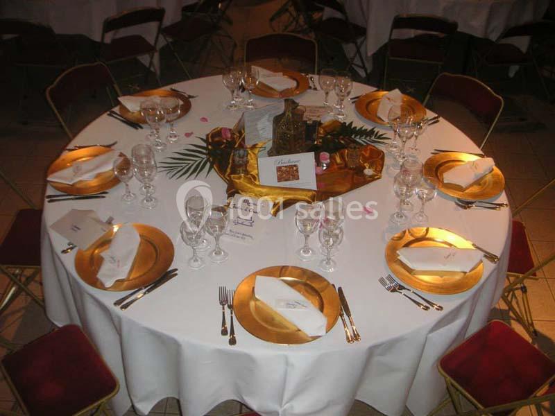 Table ronde dressée avec nappes blanches, assiettes dorées, verres, serviettes pliées et décoration centrale florale.