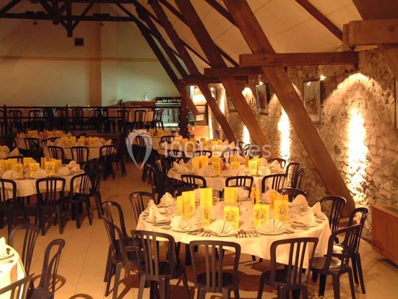 Salle de réception avec des tables rondes dressées, chaises noires et éclairage chaleureux sous une charpente en bois.