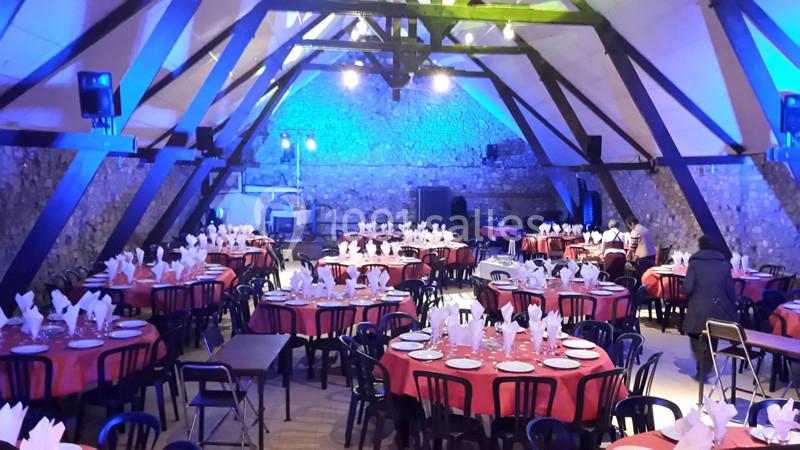Salle de réception avec des tables rondes dressées de nappes rouges, assiettes et serviettes pliées, éclairage bleu.