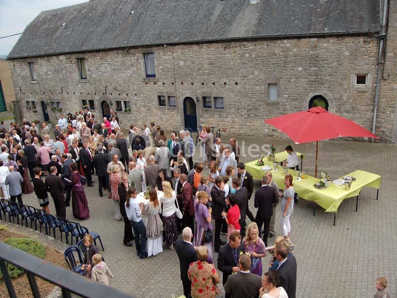 Groupe de personnes rassemblées dans une cour pavée devant un bâtiment en pierre, avec tables et parasol rouge.