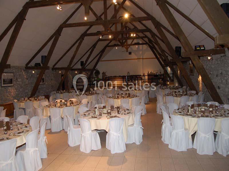 Salle de réception avec des tables rondes dressées, nappes blanches et poutres apparentes en bois.