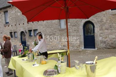 Table dressée avec nappes jaunes, seaux à champagne et verres, sous un grand parasol rouge devant un bâtiment en pierre.