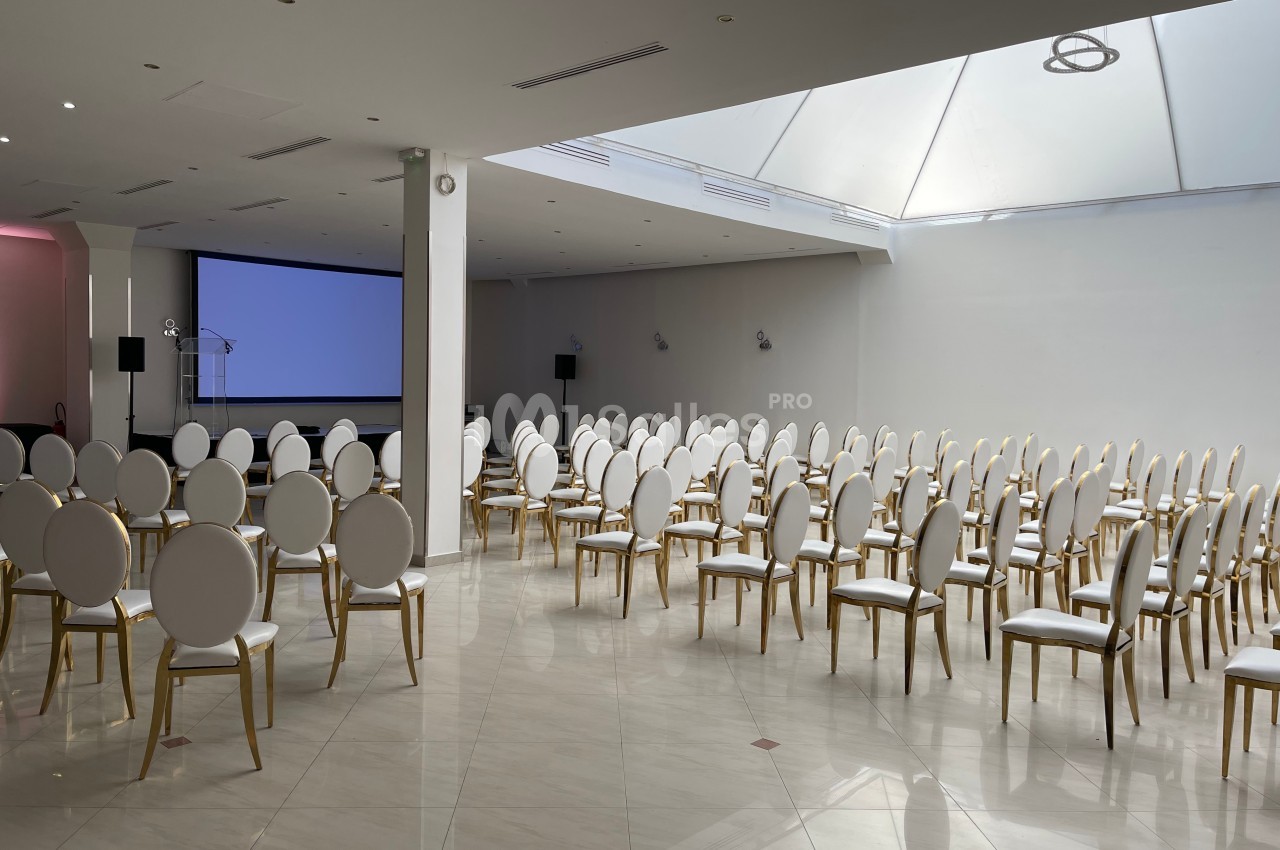 Salle de conférence lumineuse avec des rangées de chaises blanches et un écran de projection à l'arrière-plan.