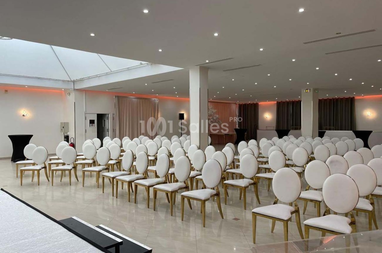 Salle lumineuse aménagée avec des rangées de chaises blanches à structure dorée, prête pour un événement.