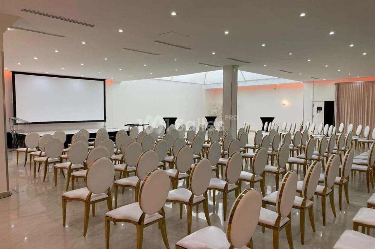 Salle de conférence lumineuse avec des rangées de chaises blanches, un écran de projection et des tables hautes noires.