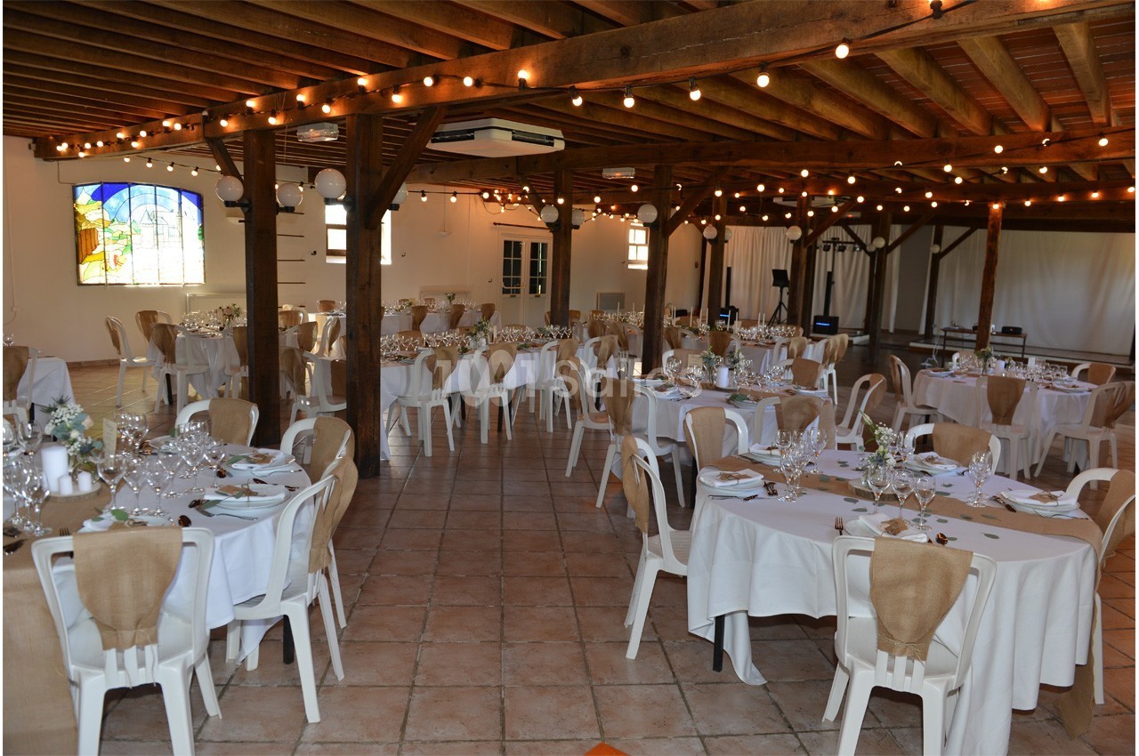 Salle de réception décorée avec des tables rondes dressées, nappes blanches et guirlandes lumineuses suspendues.