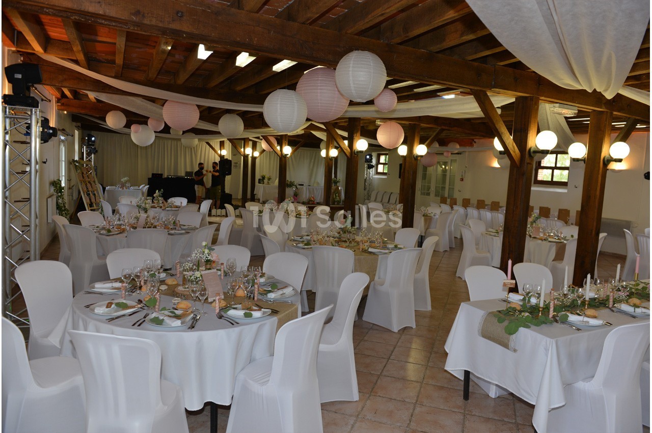 Salle de réception décorée pour un événement, avec tables rondes dressées, chaises blanches et lanternes suspendues.