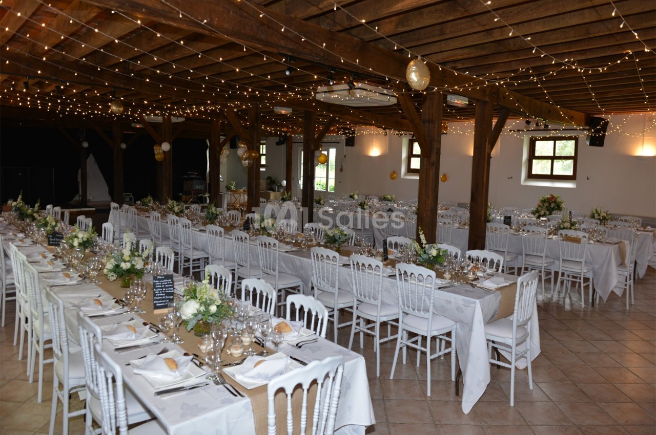 Grande salle de réception décorée avec des guirlandes lumineuses, des tables dressées et des chaises blanches.