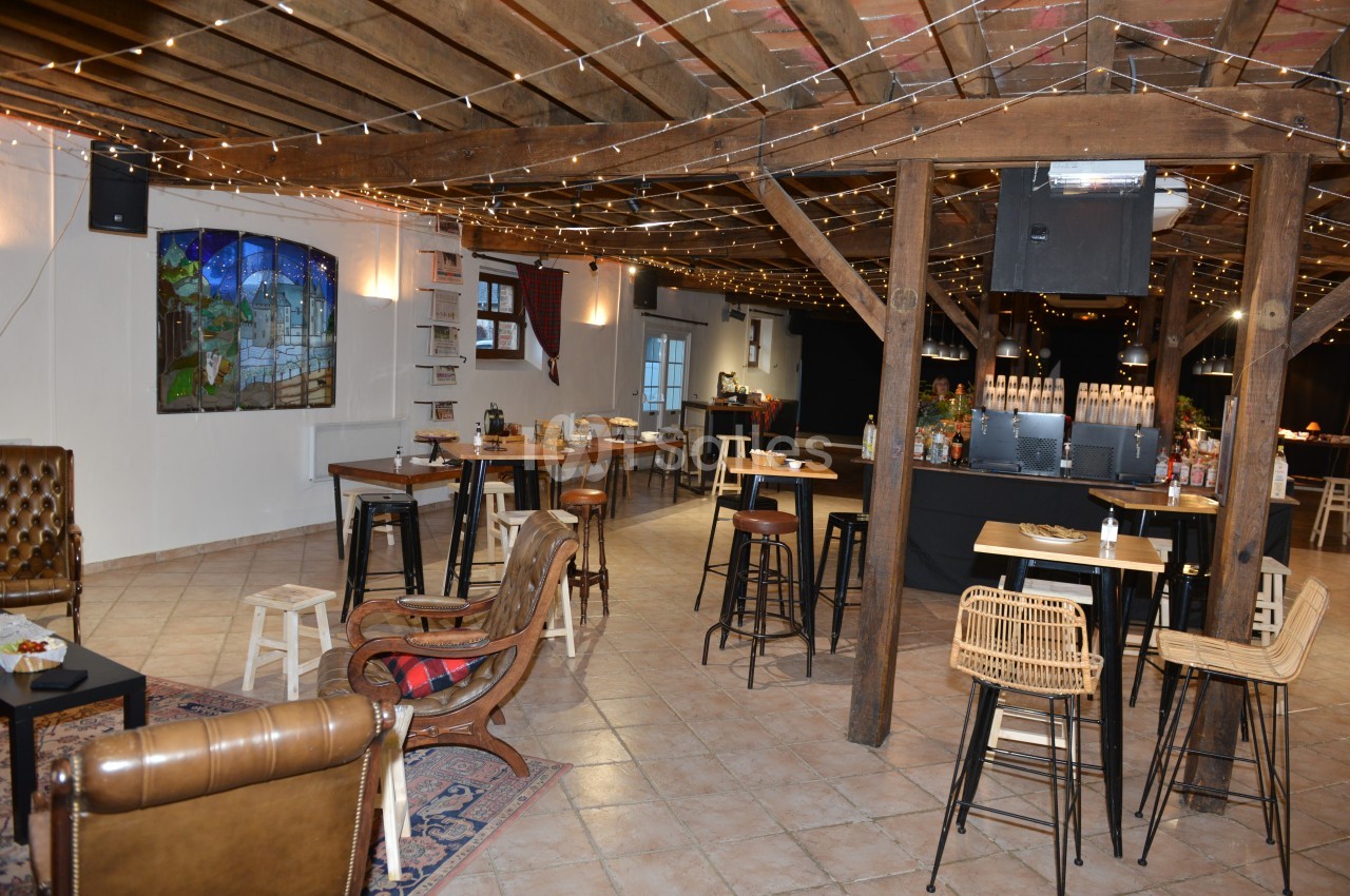 Salle aménagée avec des tables hautes, des chaises en bois et un bar, éclairée par des guirlandes lumineuses suspendues.