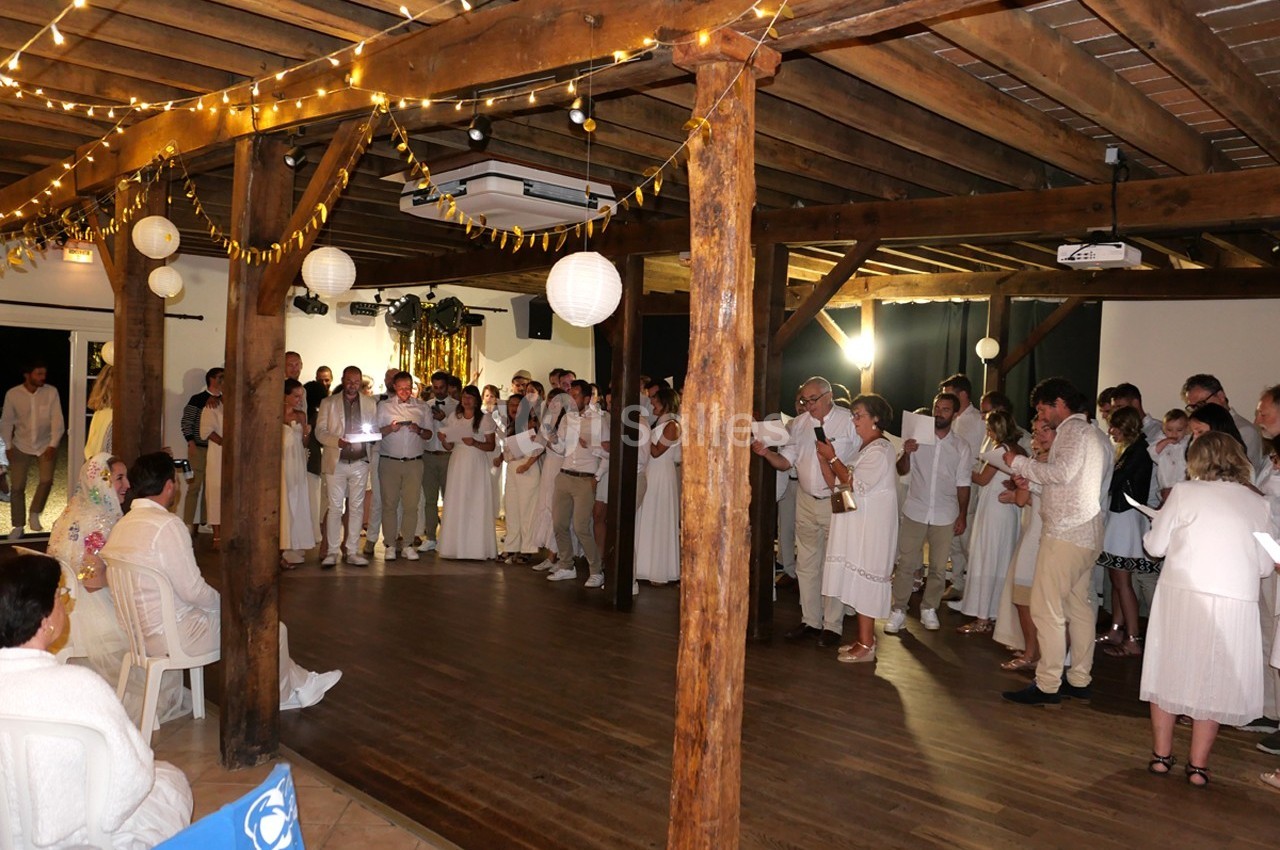 Groupe de personnes vêtues de blanc rassemblées dans une salle en bois décorée de guirlandes lumineuses et de lanternes.